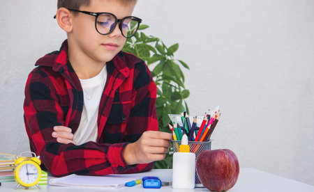 Smart young boy in glasses preparing for school, choosing pencils from the organizer on his desk