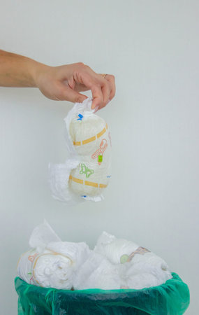 A woman's hand disposing of a used baby diaper into a trash can with a green liner.