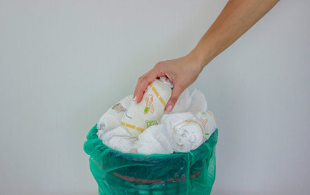 A woman's hand disposing of a used baby diaper into a trash can with a green liner.