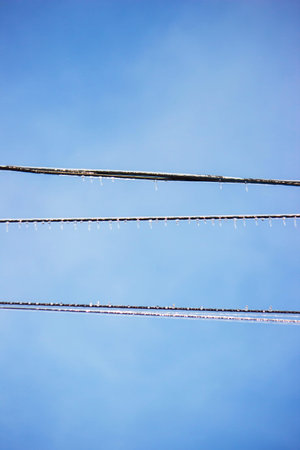 Icy icicles on electric wires against blue sky, freezing rain consequences.