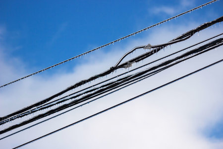 Icy icicles on electric wires against blue sky, freezing rain consequences.