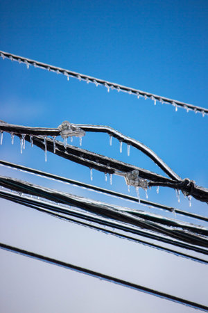 Icy icicles on electric wires against blue sky, freezing rain consequences.