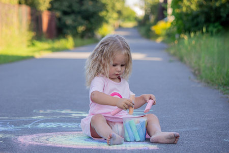 Cute little girl drawing a colorful rainbow with chalk on the asphalt road.