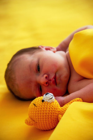 Newborn baby lying on a yellow blanket with a small knitted teddy bear toy.の写真素材