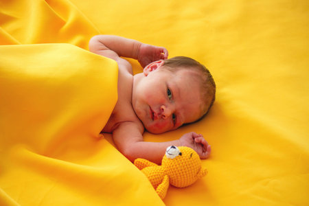 Newborn baby lying on a yellow blanket with a small knitted teddy bear toyの写真素材