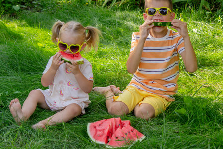 Brother and sister in sunglasses eating watermelon on the grass in summerの写真素材