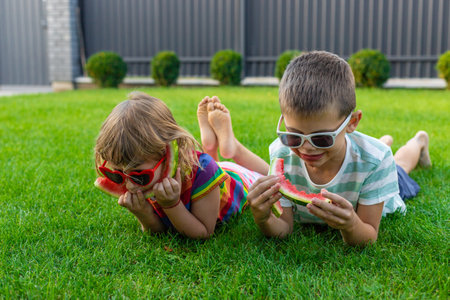 Happy children in sunglasses eating watermelon slices on green grass.の写真素材