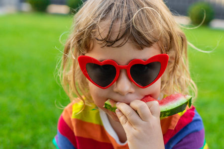 Little girl in heart-shaped sunglasses eating watermelon on green grass.の写真素材