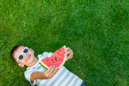 Boy in sunglasses eating a juicy watermelon slice sitting on green grass.の写真素材
