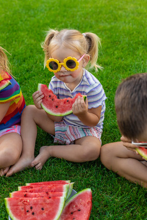 Little girl in yellow flower sunglasses eating watermelon on green lawn.の写真素材