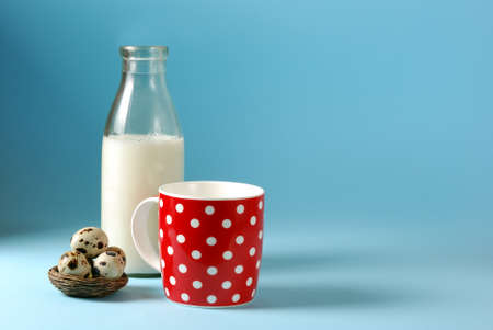 Still life with red, in polka dot, cup of milk, quail eggs, and antique bottle of milk on a blue backgroundの写真素材