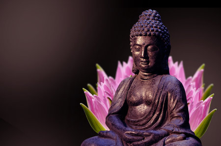 Buddha statue sitting in meditation pose against deep dark background and pink lotus flower behind.