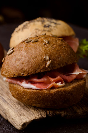 Delicious crispy burger bun with sliced ham on a vintage wooden cutting board. Dark moody food photo...