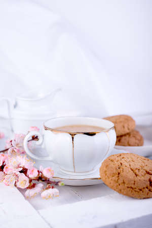 White porcelain cup of tea, milk jug, and fresh-baked oat cookies. English breakfast still-life with drink and treats and white tablecloth.の写真素材