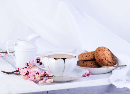 White porcelain cup of tea, milk jug, and fresh-baked oat cookies. English breakfast still-life with drink and treats and white tablecloth.の写真素材