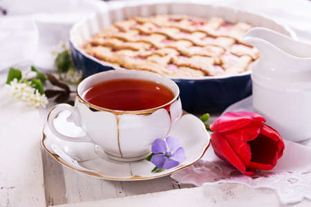 White porcelain cup of tea, and fresh-baked pie. British breakfast still-life with drink and treats, red tulip flower and white tablecloth.の写真素材