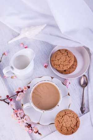 White porcelain cup of coffee or cacao, milk jug, and fresh-baked oat cookies. English breakfast still-life with drink and treats and white tablecloth.の写真素材