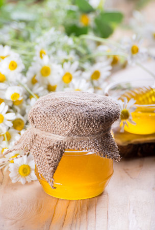 Sweet spring herbal honey in the jar and small glass bowl with a honey dipper. Composition of aromatic honey on the table with fresh chamomile flowers.の写真素材