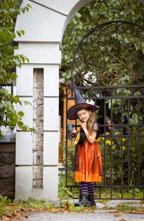 Happy little girl wearing a witch carnival costume and basket for sweets trick or treating near neighbors yard gates.の写真素材