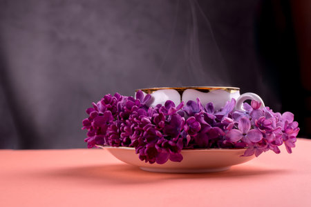Porcelain cup of tea and branch of lilac flowers on a gentle coral color background. Spring seasonal...