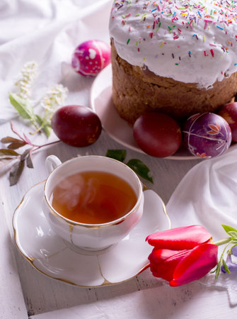 Happy Easter breakfast still life with White porcelain cup of tea, colored eggs, and fresh-baked...