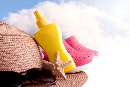 Sunscreen spray bottle and summer straw hat beach shoes and sunglasses against cloudy sky. Summer vacation banner concept.の写真素材