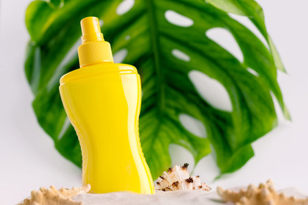 Sunscreen spray bottle on a sandy beach with different seashells against big monstera leaf...