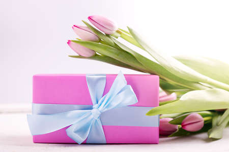 Romantic gift box tied with big pink bow and spring tulips on white wooden surface. Mother s day or Easter holiday minimalist concept.の写真素材