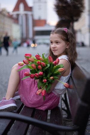 Adorable 9 years old girl with long curly hair walking through central square of the town Olesnica, Poland. Beautiful girl walking with bouquet of tulip flowers through spring European city.の写真素材