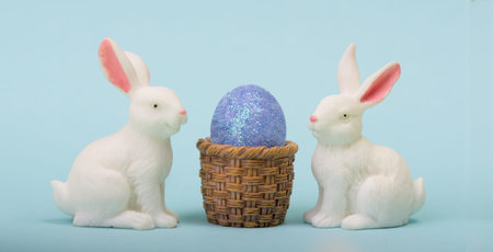 Pair of white Easter bunny figurines against blue background with Easter egg and the basket copy...