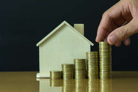 male hand putting golden coin stack growth up with wooden house in background, saving conceptの写真素材