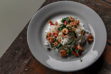 Rice topped with Stir fried thai basil with dwarf prawn served in white dish on wooden table, A...