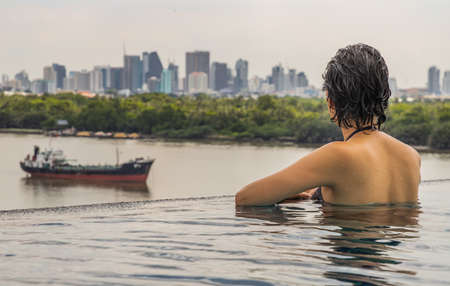 Short haired young woman relax sitting on the edge of roof top outdoor swimming pool and enjoying the city view Bangkok. Summer vacation concept.の写真素材
