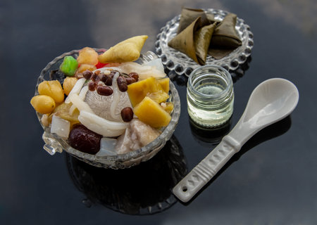 Traditional chinese iced sweet dessert is delicious here are many grains including Sweet potato balls, Grass jelly, Red beans, Ginkgo, Taro balls, lotus seeds, millet, persimmon, longan and boiled flour in Glass bowl Served with Sticky rice in banana leaf and boil sugar syrup. Selective focus.の写真素材