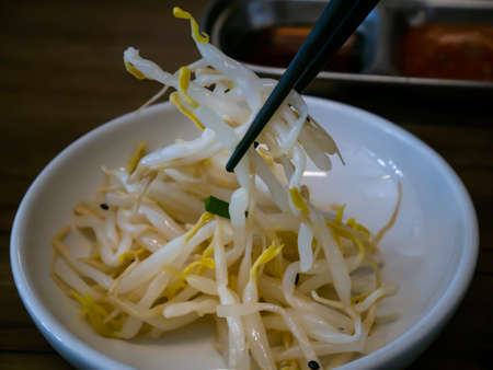 Hand using chopsticks pick Korean seasoned beansprout (Mild kongnamul muchim) in a white cup for eating. Asian side dish Pickled radish, Korean food traditional. No focus, specifically.の写真素材