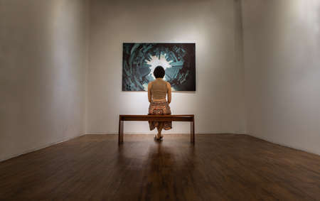 Bangkok, Thailand - 24 Feb 2021 : Back view of young asian woman sitting on bench is interested in photograph displayed on white wall at art gallery exhibition hall. Concept of fine art in photographic gallery. Selective focus.のeditorial素材