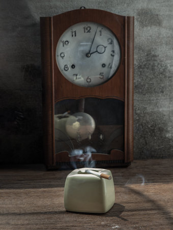 A litted cigarette on greyish green ashtray which is placed in front of a vintage wood pendulum...