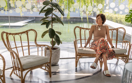 A young asian woman sitting and relax on cushion on rattan sofa  near glass window in living room. Selective focus.の写真素材