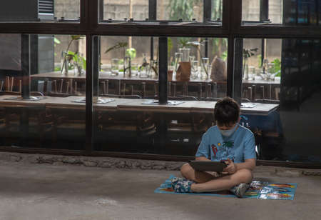 Bangkok, Thailand - 22 Jun 2021 : A boy sitting alone on the floor and watching something on Ipod outside a cafe. Copy space, Selective focus.のeditorial素材
