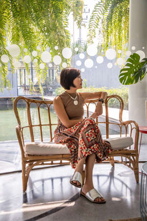A young asian woman sitting and relax on cushion on rattan sofa  near glass window in living room. Selective focus.の写真素材