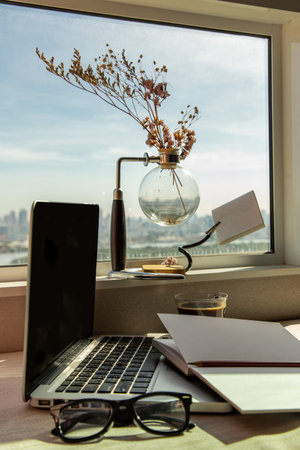 Working space with laptop, notebook, pencil, sunglasses, coffee cup, dried flower vase by the window...
