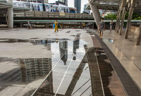 Photo reflections of bridge structure, buildings and sky above a puddle of rain water on the skywalk to skytrain station at "Chong Nonsi" Copy space, Selective focus.のeditorial素材