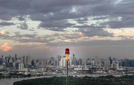 Red lantern of obstruction lights mounted on the rooftop of high rise tall building to ensure flights safety and warn the danger for the plane on the city view background at sunset. Red alarm lamp, Selective focus.の写真素材