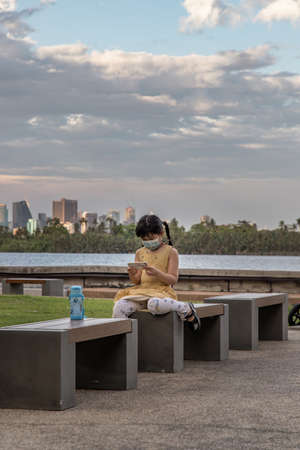 Bangkok, Thailand - 02 Feb, 2022 : A little girl wearing mask to protect covid-19 and watching something on smartphone while sitting on the black bench in the waterfront park. Wears face mask for the coronavirus pandemic. Selective focus.のeditorial素材
