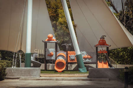 Shaded kid's playground activity tower equipment at park with a slide. Chidren's playing area outdoor, Made from metal and plastic, Under tension membrane roof on green garden background.Selective focus.の写真素材