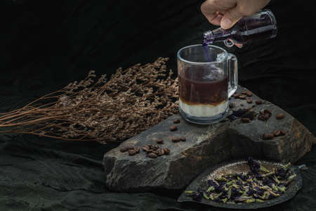 Pouring butterfly pea juice (anchan) from small glass bottle into the glass of espresso coffee and fresh milk at the bottom with roasted coffee beans and dry butterfly pea flowers nearby on dark background. Healthy detox herbal drink, space for text, Selective focus.の写真素材