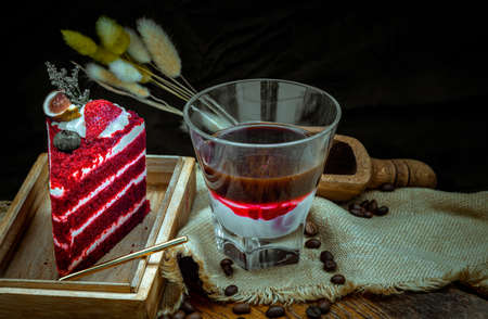 Concentrated flavored syrup mixed Coffee with fresh milk at the bottom of transparent glass serve with Piece of red velvet cake on Wooden cutting board. Coffee break, Selective focus.の写真素材