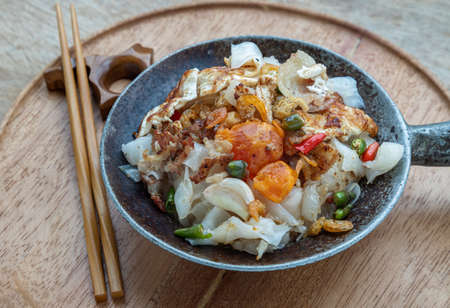 Stir-fried wide rice noodles with Bacon, Egg, Dried shrimp in small steaming iron pot and wooden chopsticks served on round wooden tray. Selective focus.の写真素材