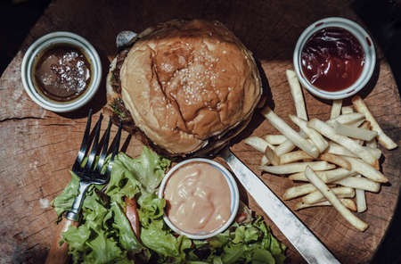Pork burger with white sesame seeds with French fries potatoes and Salad served with ketchup sauce, mayonnaise and salad dressing on wooden cutting board. The concept of delicious food, Top view, Selective Focus.の写真素材