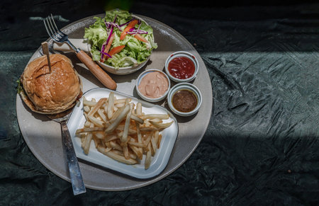 Pork burger with white sesame seeds with French fries potatoes and Salad served with ketchup sauce, mayonnaise and salad dressing on wooden cutting board. The concept of delicious food, Top view, Copy space, Selective Focus.の写真素材